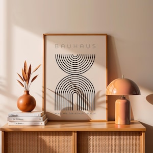 Op de afbeelding: Een ingelijste Bauhaus-kunstprint met een geometrisch ontwerp in zwart op een crèmekleurige achtergrond. Een terracotta vaas met gedroogde bladeren, boeken en een perzikkleurige lamp staan op een houten kast.