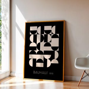 Könnte beinhalten: Ein gerahmter Bauhaus-Kunstdruck mit einem geometrischen Design in Schwarz und Off-White. Das Kunstwerk zeigt Halbkreise und Quadrate mit dem Text "BAUHAUS 1919" unten. Der Rahmen ist goldfarben, und der Druck wird in Innenräumen ausgestellt.