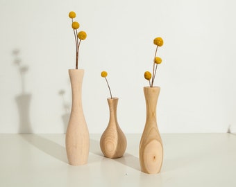 以下が含まれることがあります： 高さや形が異なる3つの木製の花瓶で、シンプルで自然なデザインです。花瓶は空ですが、1つは乾燥した黄色の花が添えられています。