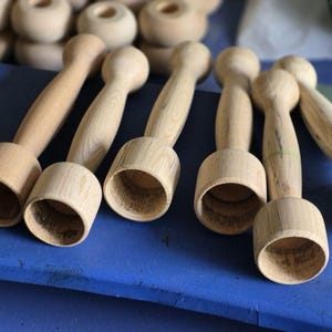 Op de afbeelding: Verschillende onafgewerkte houten items, mogelijk voor knutselen of houtbewerking, liggen op een blauw oppervlak. De items hebben een cilindrische basis en een afgeronde bovenkant, verbonden door een smallere steel. De natuurlijke houtnerf is zichtbaar.