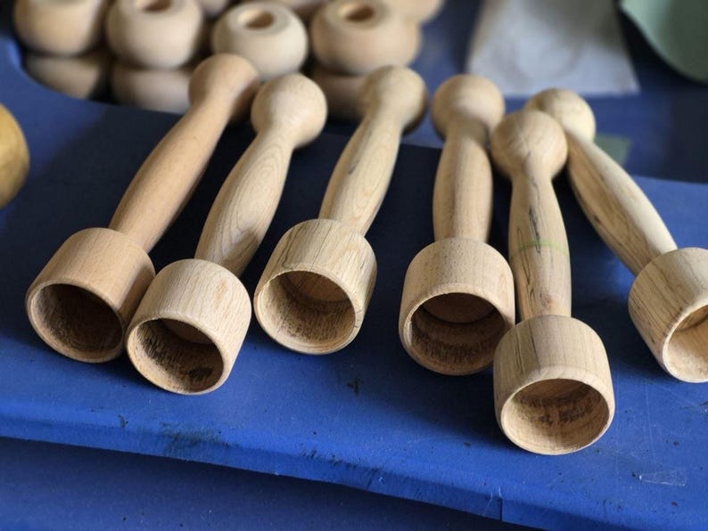 Op de afbeelding: Verschillende onafgewerkte houten items, mogelijk voor knutselen of houtbewerking, liggen op een blauw oppervlak. De items hebben een cilindrische basis en een afgeronde bovenkant, verbonden door een smallere steel. De natuurlijke houtnerf is zichtbaar.