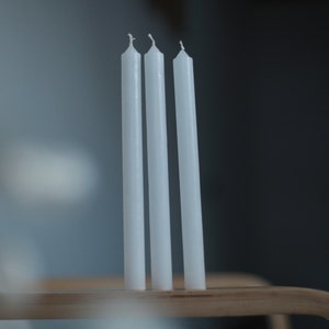 May include: Three white taper candles standing upright on a wooden surface.