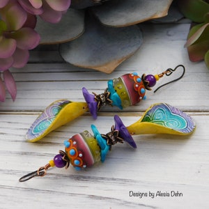May include: A pair of handmade earrings featuring colorful glass beads and polymer clay elements. The earrings have a yellow, teardrop-shaped base with a blue and purple design. The beads are multi-colored with turquoise accents and purple details.