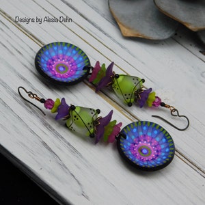 May include: Handmade earrings featuring a circular, blue and purple design with a central pink and green pattern. The earrings also include green bell-shaped beads, purple flower-shaped beads, and pink beads. The ear wires are dark metal.