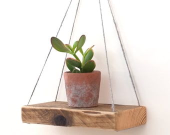 Könnte beinhalten: Ein rustikales Holzregal mit einem Hängeseilsystem, das eine kleine Topfpflanze mit grünen Blättern hält. Das Regal ist aus recyceltem Holz gefertigt und hat eine natürliche, verwitterte Oberfläche.