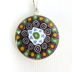 以下が含まれることがあります： 茶色、青、緑、黄色、赤の色合いで、複雑でカラフルなデザインが施された丸いペンダント。ペンダントはムラーノガラス製で、銀で縁取られています。