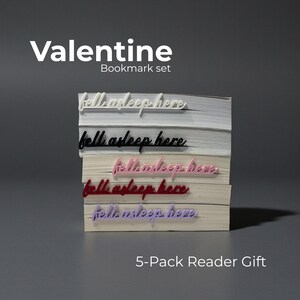 Pode incluir: Um conjunto de marcadores de livros de Dia dos Namorados com cinco marcadores nas cores branca, preta, rosa, vermelha e roxa. Cada marcador, com a inscrição cursiva "fell asleep here", está sobre uma pilha de livros. O texto "Valentine Bookmark set" e "5-Pack Reader Gift" também é visível.