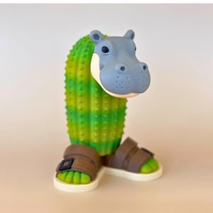 Puede incluir: Una figura impresa en 3D con una cabeza de hipopótamo gris unida a un cuerpo de cactus espinoso con rayas verdes y amarillas. La figura tiene pies verdes con sandalias marrones y hebillas negras, sobre una base blanca plana.