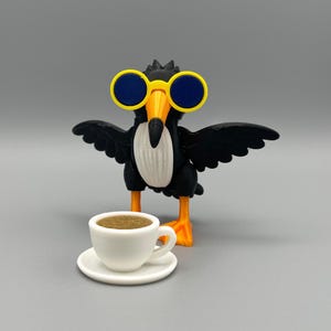 Puede incluir: Una figura de pájaro negro con pecho blanco, pico y patas naranjas, que lleva gafas de sol redondas con montura amarilla y cristales azules, con las alas extendidas. Frente al pájaro hay una pequeña taza de café blanca con plato.