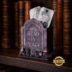 Puede incluir: Un sujetalibros con forma de lápida de color morado oscuro con el texto READ IN PEACE en relieve en la parte frontal. La base está decorada con calaveras y rosas esculpidas. Dentro hay dos tarjetas rectangulares blancas con una máscara de hockey.