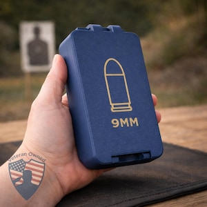 May include: A navy blue 9MM ammunition case with a gold bullet graphic. The case is rectangular with a hinged lid. The text "9MM" is printed in gold below the bullet graphic. The case is held in a hand.