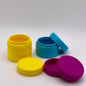 Puede incluir: Tres pequeños recipientes redondos en amarillo, turquesa y magenta. Cada recipiente tiene un patrón geométrico texturizado en los lados y una tapa a juego. Las tapas se colocan junto a los recipientes, con una tapa abierta.