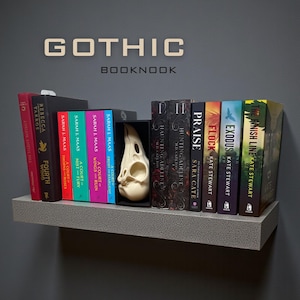 May include: A gray shelf displaying a collection of books with colorful spines. The books include titles like "Fourth Wing," "A Court of Thorns and Roses," and "Exodus." A decorative skull is placed within a black box. The word "GOTHIC BOOKNOOK" is displayed above.