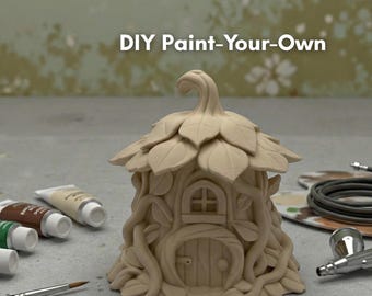 Könnte beinhalten: Ein ungefertigtes, beiges DIY-Feenhaus mit blattförmigem Dach und weinartigen Details. Der Text "DIY Paint-Your-Own" steht über dem Haus. Farbtuben und Pinsel im Vordergrund deuten auf ein Bastelprojekt hin.