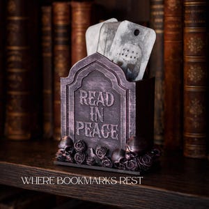 May include: A tombstone-shaped bookmark holder with the words "READ IN PEACE" in white lettering. The holder is dark gray with skulls and roses at the base. The phrase "WHERE BOOKMARKS REST" is at the bottom. Several bookmarks are inside.