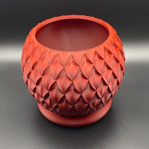 Puede incluir: Una maceta esférica roja con un patrón texturizado en forma de escamas. La maceta tiene una abertura ancha y se asienta sobre una base pequeña y redonda. El diseño general recuerda a un huevo de dragón. Adecuado para plantas pequeñas o suculentas.