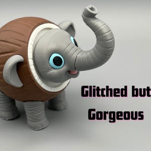 K&ouml;nnte beinhalten: Eine 3D-gedruckte Figur eines grauen Elefanten mit einem braunen, kokosnussf&ouml;rmigen K&ouml;rper, gro&szlig;en blauen Augen und einem erhobenen R&uuml;ssel. Der Text Glitched but Gorgeous ist in schwarzen Buchstaben rechts neben der Elefantenfigur aufgedruckt.