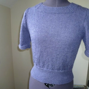 May include: A short-sleeved, knitted sweater in a light purple color. The sweater has a round neckline and a slightly fitted silhouette.
