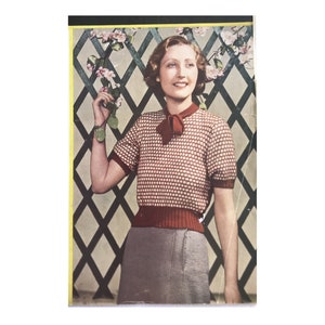 May include: A woman wearing a red and white knitted sweater with a bow at the neck. She is standing in front of a trellis with pink flowers.