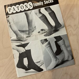 May include: A monochrome advertisement for Patons Family Socks, featuring various sock styles. The image is divided into four sections, displaying different designs, including patterned and ribbed socks. The text "PATONS Family Socks" is visible at the top.