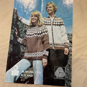 Könnte beinhalten: Vintage Zeitschriften-Cover mit einem Mann und einer Frau in gemusterten Strickpullovern. Die Frau trägt einen braunen Pullover mit weißen und braunen geometrischen Mustern. Der Mann trägt einen weißen Pullover mit braunen Rautenmustern. Das Magazin hat den Text "ÁLAFOSS LTD. ICELAND" und "PURE NEW WOOL".