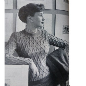 May include: A black and white photo of a woman wearing a cable knit sweater. She is sitting with her arm resting on a surface and looking to the right.