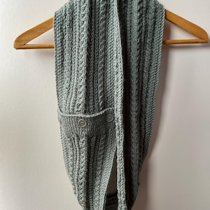 Könnte beinhalten: Ein salbeigrüner gestrickter Infinity-Schal mit vertikalem Zopfmuster und einer dekorativen Lasche, die mit zwei passenden runden Knöpfen befestigt ist. Der Schal hängt an einem Holzkleiderbügel und zeigt sein strukturiertes Design und die sanfte Farbe.