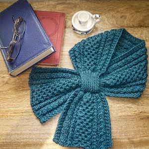 May include: A teal knitted scarf with a textured pattern, displayed on a wooden surface. The scarf is folded in a bow shape. A blue book, a red book, a pair of glasses, and a small ceramic cup are in the background.