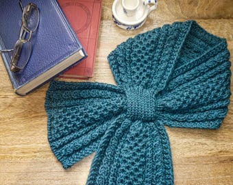 May include: A teal knitted scarf with a textured pattern, lying on a wooden surface. The scarf is folded in a bow shape. A blue book, a red book, a pair of glasses, and a small ceramic cup are in the background.