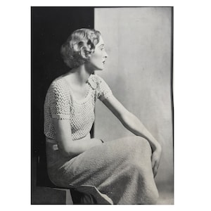 May include: A black and white photograph of a woman wearing a crocheted top and skirt. She is sitting with her right arm resting on her lap and her left arm resting on her knee. The background is a light gray wall.