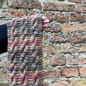 May include: A cable-knit scarf in shades of brown, pink, and beige. The scarf is hand-knitted, with a textured pattern. The yarn appears soft and warm. The background is a weathered brick wall. The scarf is a cozy accessory for colder weather.