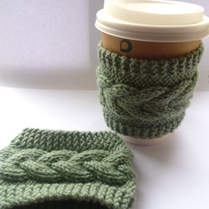 May include: A green knitted coffee cup cozy with a cable knit pattern. The cozy is on a white surface and a brown paper coffee cup with a white lid is next to it.