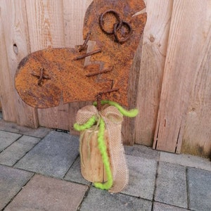 Könnte beinhalten: Eine rostige Metallherzsculptur mit einer Schleife aus Sackleinen und grünem, flauschigem Besatz, montiert auf einem Holzscheit. Das Herz ist gebrochen und mit Draht zusammengenäht.