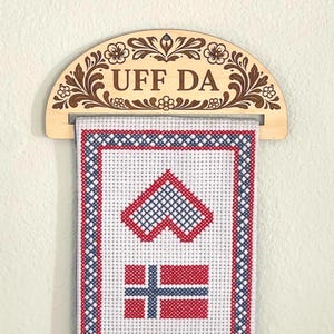 May include: Wooden wall hanging with the text "UFF DA" and floral carvings. The hanging features a white fabric with red and blue cross-stitch designs of hearts and a Norwegian flag.
