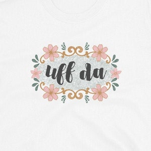 May include: White t-shirt with the phrase "uff da" in black script, surrounded by a floral design with pink flowers and green leaves. The design is set against a textured background.