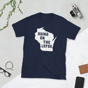 May include: Navy blue t-shirt featuring a white outline of Wisconsin with the text "BRING ON THE LEFSE" in a distressed font. The shirt is made of a soft material and is suitable for casual wear. A camera, glasses, and a wallet are in the background.