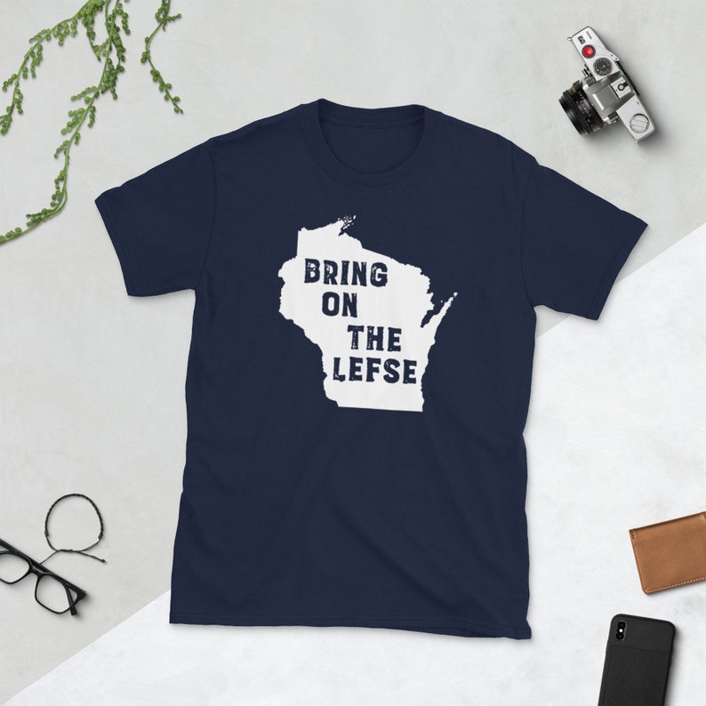May include: Navy blue t-shirt featuring a white outline of Wisconsin with the text "BRING ON THE LEFSE" in a distressed font. The shirt is made of a soft material and is suitable for casual wear. A camera, glasses, and a wallet are in the background.