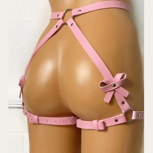 May include: Pink leather harness with a silver ring and bow accents. The harness has adjustable straps and is designed to be worn around the waist and thighs.