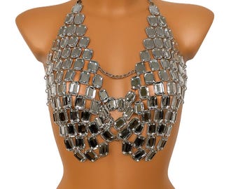 May include: A silver-colored body chain top composed of numerous rectangular, reflective pieces. The halter-style top is connected by a delicate chain around the neck and across the chest, creating a shimmering, geometric design. The top is displayed on a mannequin.
