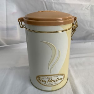 May include: A cylindrical Tim Hortons coffee canister with a tan lid and gold-coloured accents. The canister is white with a cream-coloured design and the words "ALWAYS FRESH" and "TOUJOURS FRAIS" printed on the front.