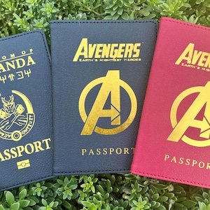 May include: Three passport covers, one black with gold lettering that says "Kingdom of Wakanda" and "Wakanda" in a different font, one black with gold lettering that says "Avengers Earth's Mightiest Heroes" and a gold Avengers logo, and one red with gold lettering that says "Avengers Earth's Mightiest Heroes" and a gold Avengers logo.
