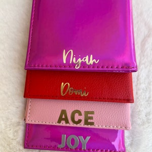 May include: Four colorful wallets stacked on top of each other. The top wallet is a bright pink with the name "Nijah" written in gold letters. The second wallet is red with the name "Dom" written in gold letters. The third wallet is pink with the word "ACE" written in gold letters. The bottom wallet is a bright purple with the word "JOY" written in gold letters.
