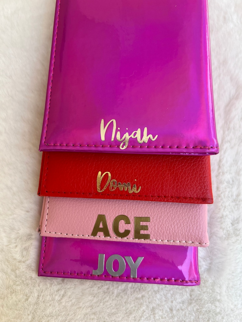 May include: Four colorful wallets stacked on top of each other. The top wallet is a bright pink with the name "Nijah" written in gold letters. The second wallet is red with the name "Dom" written in gold letters. The third wallet is pink with the word "ACE" written in gold letters. The bottom wallet is a bright purple with the word "JOY" written in gold letters.