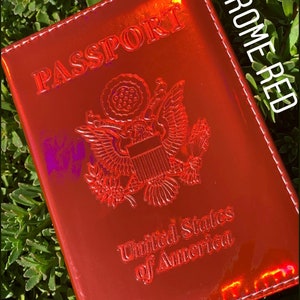 May include: A red passport holder with a chrome finish. The holder has the words "PASSPORT" and "United States of America" embossed on the front. The holder is labeled "CHROME RED".