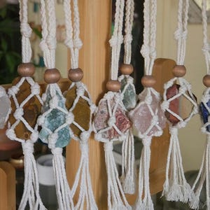 May include: A collection of macrame hangers featuring off-white cotton cord knotted in a net pattern around various raw gemstones. Each hanger includes a smooth wooden bead above the stone and ends in a fringe tassel at the bottom.