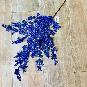 May include: A single stem of artificial berries in a vibrant blue hue. The berries are clustered along the stem, creating a full and textured appearance. The stem is brown and the berries are a deep, saturated blue. Ideal for home decor.