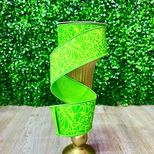 May include: A green ribbon with a floral pattern wrapped around a gold vase. The vase has a round base and a tall, slender neck.