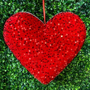 May include: A large, red heart-shaped ornament covered in sequins, hanging against a backdrop of green foliage. The ornament has a silver trim and a red ribbon for hanging. A festive decoration.