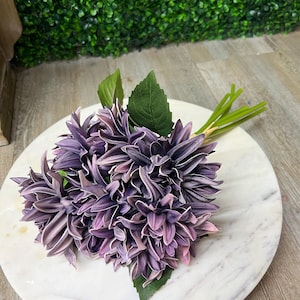 May include: A bouquet of artificial purple dahlias with green leaves and stems. The flowers are arranged on a white marble-like surface, with a green hedge background. The dahlia petals have a gradient of purple and pink hues.