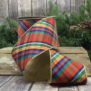 May include: A wide spool of wired ribbon featuring a vibrant plaid pattern in red, green, blue, and yellow. The ribbon has a metallic gold underside and gold-trimmed edges, with the fabric wrapped around a central core in a decorative loop.